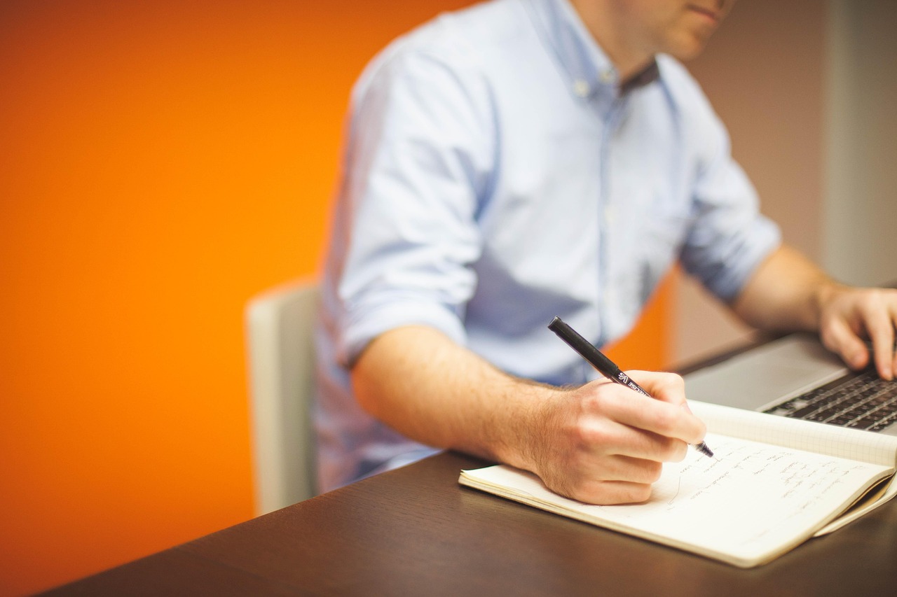 An employee in an office writing in a notebook next to a laptop